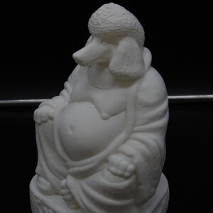 May include: A white sculpture of a seated Buddha with the head of a poodle. The figure is in a relaxed pose, with a large belly and draped robes. The base has a decorative, swirling pattern. The background is black.