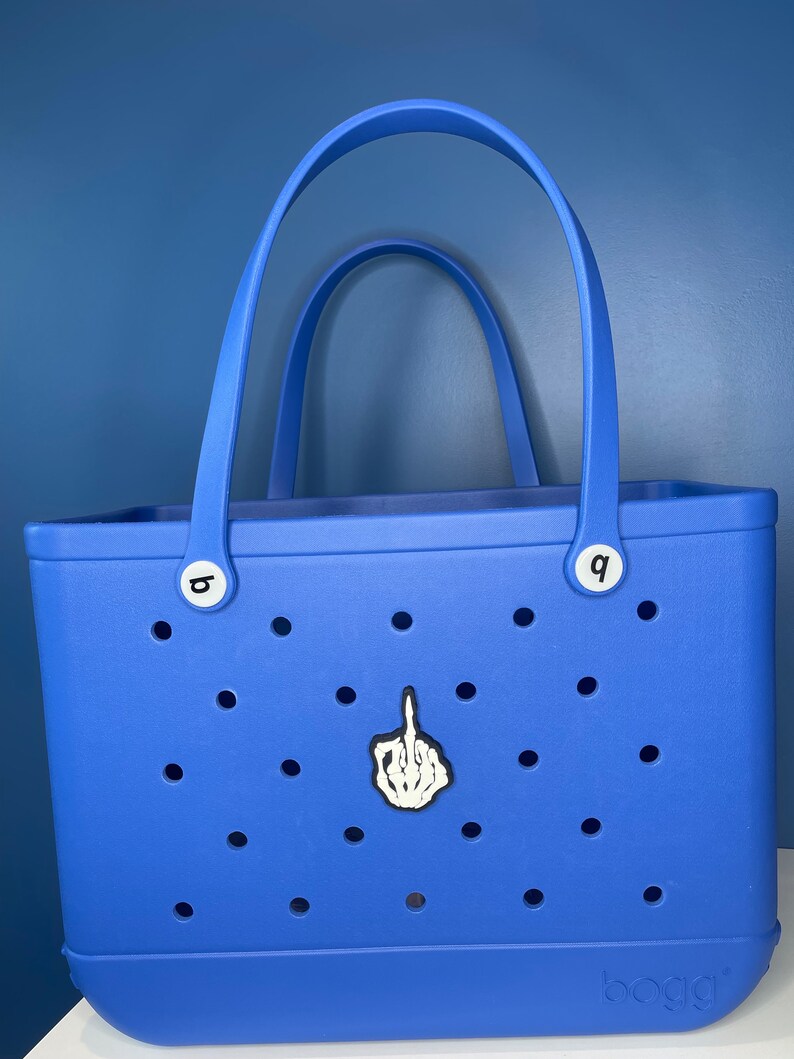 Op de afbeelding: Een blauwe tote bag met een witte skelethand die de middelvinger opsteekt. De tas heeft een wit "boga" logo aan de onderkant en witte letterknoppen aan de zijkanten.