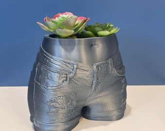 May include: A gray, 3D printed planter shaped like a pair of denim shorts with a distressed look. The planter is filled with a green succulent plant.