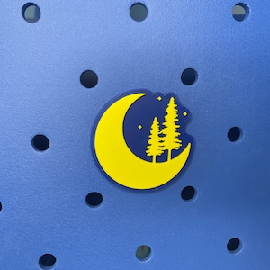 May include: A yellow crescent moon with a dark blue interior, featuring two dark blue pine trees and white stars. The moon is on a blue background with a pattern of small holes.