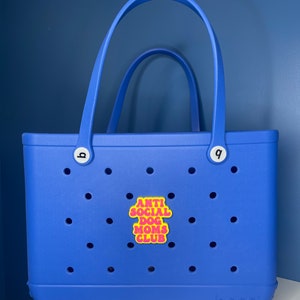 May include: A blue tote bag with a yellow patch that says "Anti Social Dog Moms Club" in pink letters. The bag has a white handle and a white logo that says "BOGA" on the bottom.