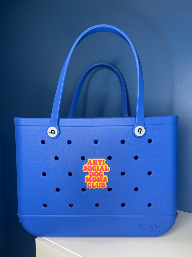 May include: A blue tote bag with a yellow patch that says "Anti Social Dog Moms Club" in pink letters. The bag has a white handle and a white logo that says "BOGA" on the bottom.