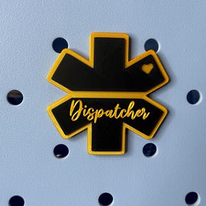May include: A black and yellow star-shaped patch with the word "Dispatcher" written in cursive on it. There is a small heart in the top right corner of the patch.