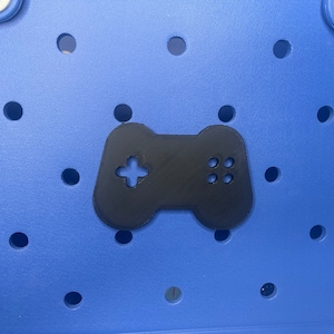 May include: A black plastic video game controller with a cross-shaped button and four round buttons. The controller is on a blue background with holes.