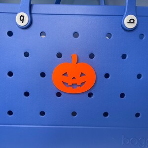 May include: A blue tote bag with a pumpkin cutout on the front. The pumpkin is orange with a black face and is outlined in white. The bag has a white handle with a white letter "q" on each handle.