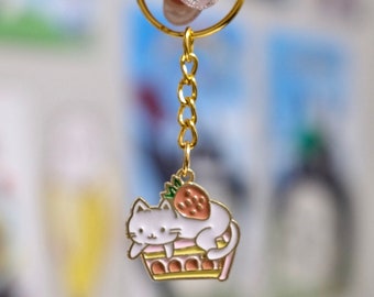 May include: Gold-colored keychain with a charm featuring a white cat resting on a pink and yellow cake, topped with a strawberry. The keychain is held by a gold ring and chain.