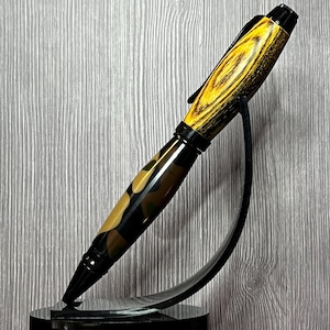 May include: A handcrafted pen with a black tip and clip, featuring a camouflage pattern in shades of brown, tan, and black. The pen's top section has a wood grain design in yellow and brown. The pen is displayed on a black stand.