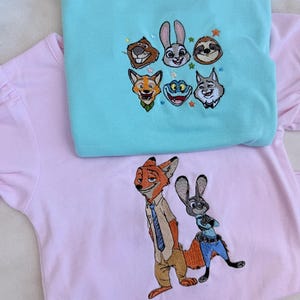 May include: Two embroidered t-shirts. One is light blue with embroidered cartoon animal faces. The other is pink with an embroidered fox and rabbit character. Both shirts have short sleeves.