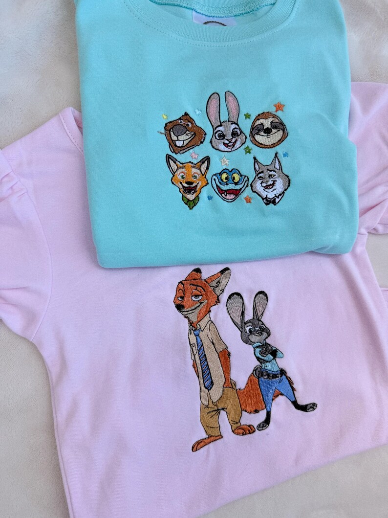 May include: Two embroidered t-shirts. One is light blue with embroidered cartoon animal faces. The other is pink with an embroidered fox and rabbit character. Both shirts have short sleeves.