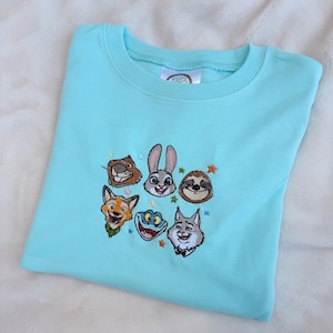 A light blue t-shirt featuring embroidered cartoon animal faces. The design includes a rabbit, fox, sloth, and other characters, with small star accents. The shirt is made of soft material. Zootopia monogram shirt