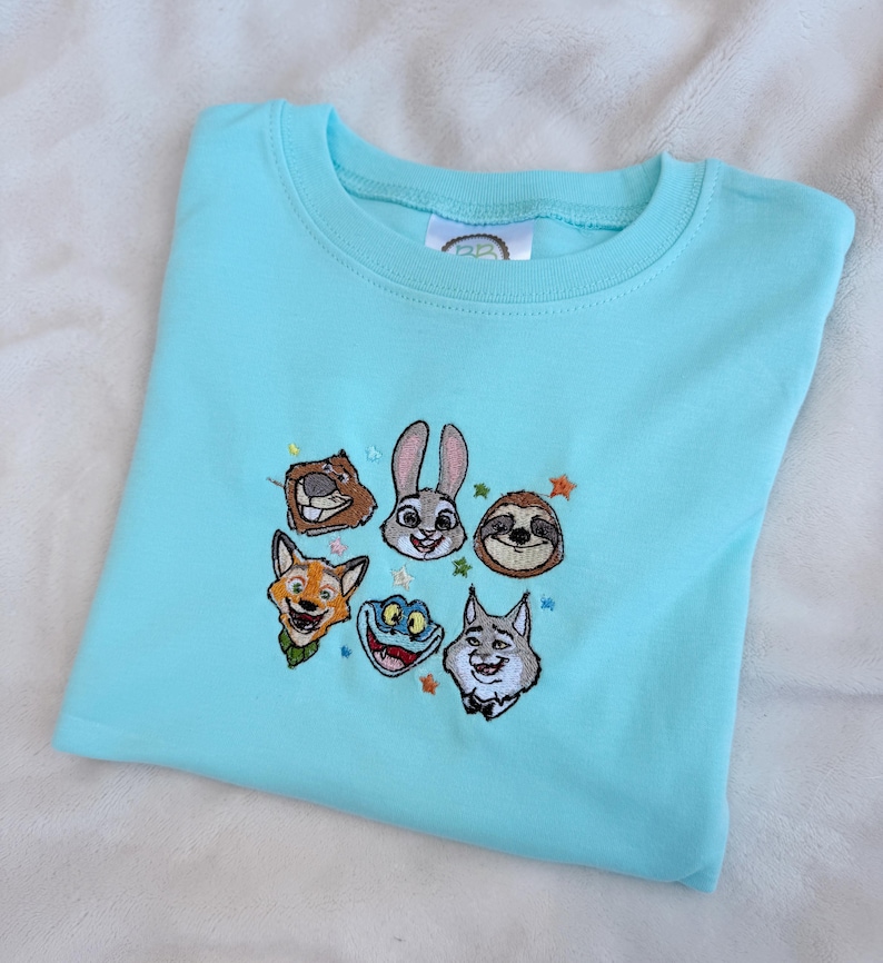 A light blue t-shirt featuring embroidered cartoon animal faces. The design includes a rabbit, fox, sloth, and other characters, with small star accents. The shirt is made of soft material. Zootopia monogram shirt