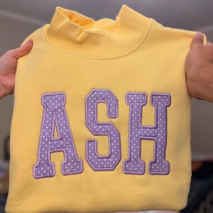 May include: A bright yellow mock neck sweatshirt featuring large, block-style letters that spell out ASH. The letters are purple with a white polka dot pattern and are stitched onto the front of the garment with a visible embroidered border.
