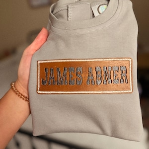 Peut inclure: Un t-shirt à manches longues gris clair avec un écusson rectangulaire en cuir marron au centre de la poitrine. L'écusson est brodé du nom JAMES ABNER en fil gris foncé et est entouré d'une bordure cousue blanche.