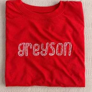 May include: A vibrant red t-shirt featuring the name "Greyson" embroidered in white, block letters. The shirt is folded, displaying the name clearly. The material looks soft and comfortable.