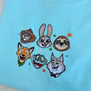 May include: A light blue sweatshirt featuring embroidered cartoon animal faces, including a beaver, rabbit, sloth, fox, frog, and wolf. The design is surrounded by small star-shaped accents. The sweatshirt has a crew neck and a soft, comfortable appearance.