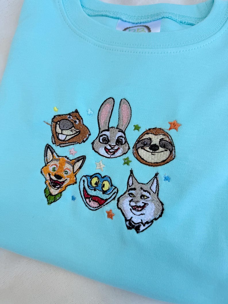 May include: A light blue sweatshirt featuring embroidered cartoon animal faces, including a beaver, rabbit, sloth, fox, frog, and wolf. The design is surrounded by small star-shaped accents. The sweatshirt has a crew neck and a soft, comfortable appearance.