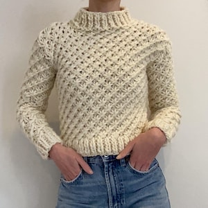May include: A cream-coloured, hand-knitted sweater with a high, ribbed collar. The sweater has a textured, diagonal stitch pattern and long sleeves. The cuffs and hem are ribbed. The sweater is paired with blue jeans.
