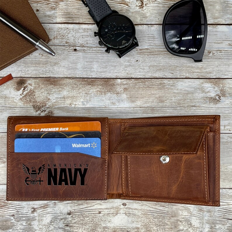May include: A brown leather wallet with a flap closure and a snap button. The wallet has a card slot and a pocket for cash. The wallet is open and has a blue and white credit card inside. The wallet has an engraved design of an eagle and the words "America's Navy".