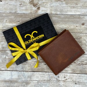 May include: A brown leather wallet with the name "Ralph Taylor" embossed on the front. The wallet is in a black gift box with a yellow ribbon and a gold logo that says "Oldenhorn expert in handcraft".