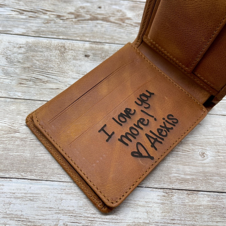 Op de afbeelding: Open, bruin leren portemonnee met de inscriptie "I love you more! Alexis" en een hartsymbool. De portemonnee is open, met kaartsleuven en een biljettenvak. Het leer heeft een verweerde uitstraling.