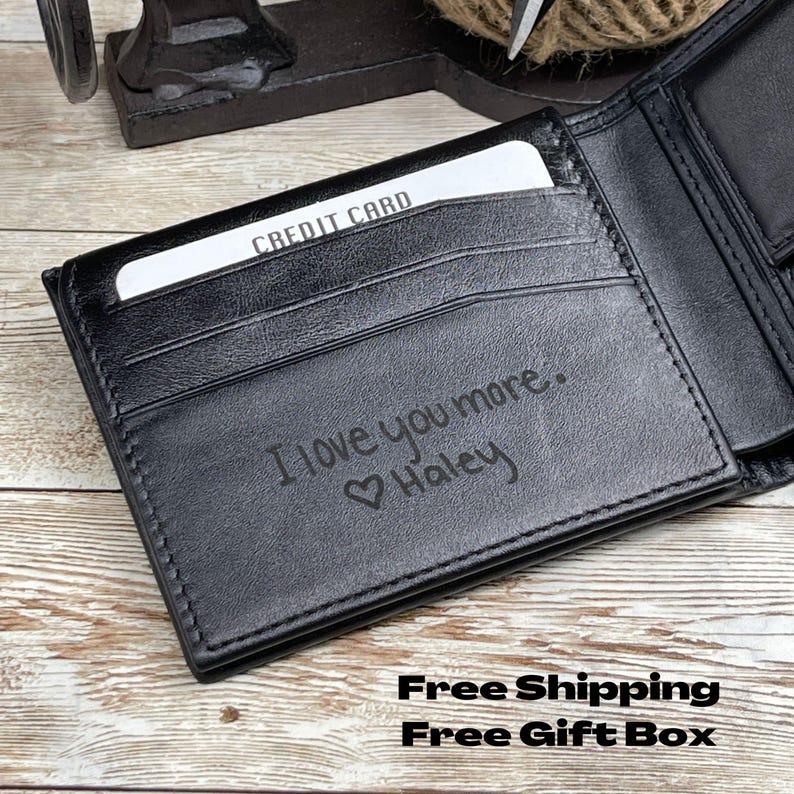 Op de afbeelding: Zwart leren portemonnee met een creditcard in een gleuf. De open portemonnee toont de gegraveerde tekst "I love you more. ❤️ Haley". De portemonnee ligt op een houten oppervlak.