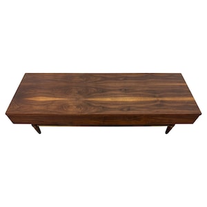 May include: A rectangular, mid-century modern coffee table made of dark walnut wood. The table has a rich, natural wood grain pattern and is supported by four angled legs. The design is simple and elegant, suitable for a living room.