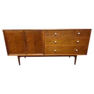 May include: A mid-century modern wooden credenza with a warm brown finish. It features two cabinet doors on the left, three drawers with white knobs on the right, and tapered legs. The credenza is a long, low piece of furniture.