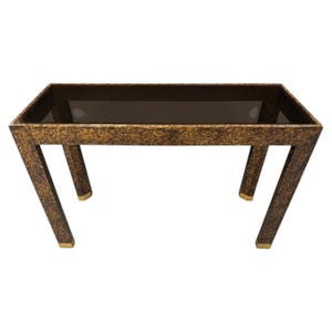 May include: A rectangular console table featuring a dark brown and gold mottled finish on the frame. The tabletop is inset with a dark, semi-transparent glass surface. The four legs are straight and finished with small gold-toned metal feet.