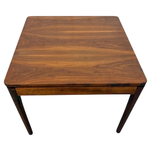May include: A square wooden side table featuring a rich, dark brown grain pattern on the flat top surface. The table has four tapered legs and a horizontal apron piece connecting the legs beneath the tabletop, showcasing a smooth finish.