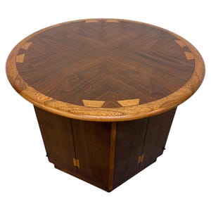 May include: A mid-century modern round coffee table. The table features a dark brown wood top with a lighter wood border and decorative inlays. The base is a dark brown, geometric shape with cabinet doors.