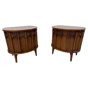 May include: Two matching mid-century modern wooden nightstands. Each has a half-moon shaped top and a curved front with decorative cutouts. The nightstands are supported by three tapered legs and are finished in a warm brown tone.