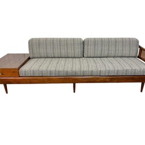 May include: Mid-century modern sofa with a built-in side table. The sofa features a wooden frame and legs, with a gray and white striped cushion. The side table has a drawer and a dark wood finish. The armrest has a woven cane design.