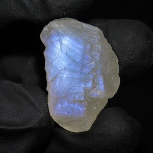 May include: A raw, uncut moonstone gemstone with a pearly white base and iridescent blue flashes. The natural stone has a rough, textured surface and is set against a dark background.