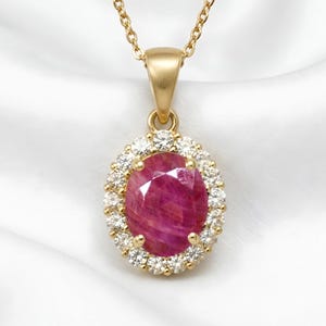 May include: A gold pendant necklace featuring a large, oval-cut, deep pinkish-red gemstone at the centre. The central stone is surrounded by a halo of small, round, clear white diamonds. The pendant hangs from a delicate gold chain link necklace.