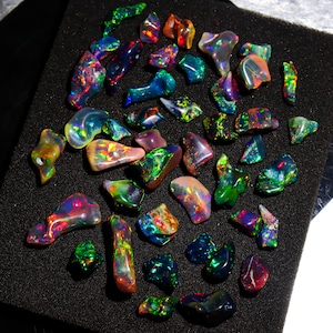May include: A collection of polished opal gemstones in various shapes and sizes. The opals display a vibrant play of color, including green, blue, red, and yellow hues. The gemstones are arranged on a black surface.