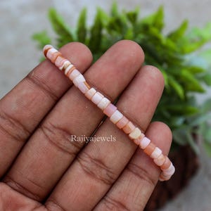 Puede incluir: Una delicada pulsera compuesta por pequeñas cuentas cuadradas de color rosa y melocotón. Las cuentas están ensartadas, creando un accesorio sutil y elegante. La pulsera se sostiene en una mano, con el texto "Rajiyajewels" visible.