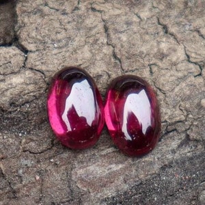 Könnte beinhalten: Zwei ovale, tiefrosa Edelsteine mit polierter, glänzender Oberfläche. Die Edelsteine reflektieren das Licht und erzeugen helle Glanzlichter. Sie liegen auf einer strukturierten, braunen Holzoberfläche, die ihre lebendige Farbe und Klarheit hervorhebt.