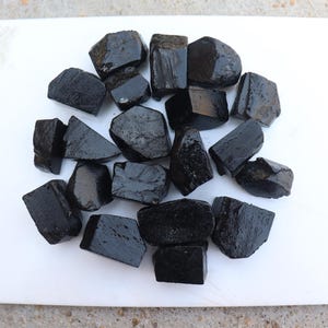 May include: A collection of irregularly shaped, glossy black tourmaline stones arranged on a white cutting board. The stones vary in size and have a dark, reflective surface, suggesting a polished finish.