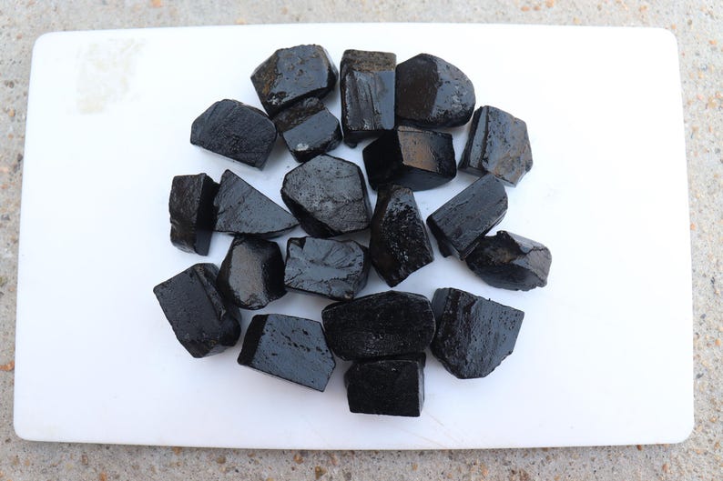 May include: A collection of irregularly shaped, glossy black tourmaline stones arranged on a white cutting board. The stones vary in size and have a dark, reflective surface, suggesting a polished finish.