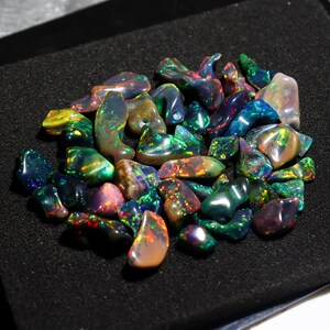 May include: A collection of polished opal gemstones in various shapes and sizes. The opals display a vibrant play of color, including green, blue, red, and yellow hues. The gemstones are arranged on a black surface, showcasing their iridescent qualities.