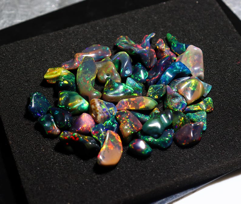 May include: A collection of polished opal gemstones in various shapes and sizes. The opals display a vibrant play of color, including green, blue, red, and yellow hues. The gemstones are arranged on a black surface, showcasing their iridescent qualities.