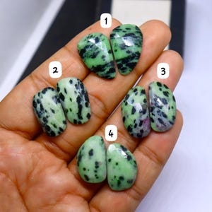 May include: Four pairs of polished zoisite gemstones. The stones are a light green color with black and dark red inclusions. The gemstones are teardrop and oval shaped. The gemstones are held in a person's hand.