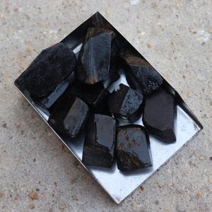 May include: A collection of raw, black tourmaline crystals in a small, square, silver metal tray. The crystals have a dark, glossy appearance and varying shapes. The tray sits on a rough, gray concrete surface.