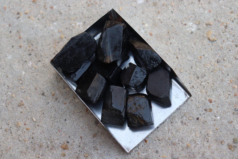 May include: A collection of raw, black tourmaline crystals in a small, square, silver metal tray. The crystals have a dark, glossy appearance and varying shapes. The tray sits on a rough, gray concrete surface.