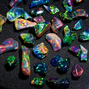 May include: A collection of polished opal gemstones in various shapes and sizes. The opals display a vibrant play of color, with flashes of green, blue, red, and yellow. The gemstones are arranged on a black surface.