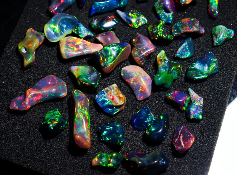May include: A collection of polished opal gemstones in various shapes and sizes. The opals display a vibrant play of color, with flashes of green, blue, red, and yellow. The gemstones are arranged on a black surface.