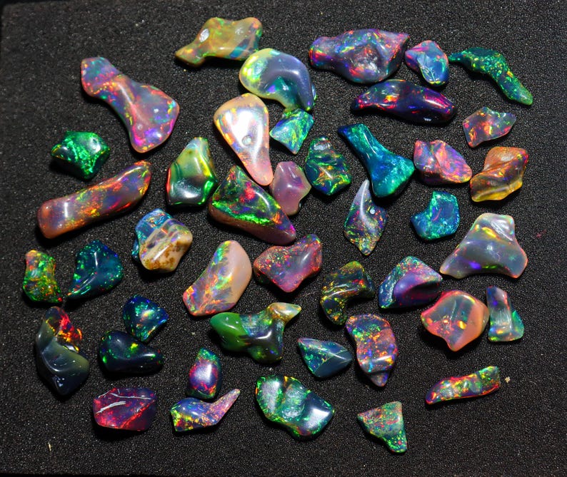 May include: A collection of polished opal gemstones in various shapes and sizes. The opals display a vibrant play of color, including shades of green, blue, purple, orange, and yellow. The gemstones are arranged on a black surface, showcasing their iridescent qualities.