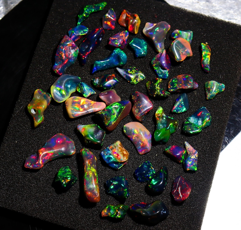 May include: A collection of polished opal gemstones in various shapes and sizes. The opals display a vibrant play of color, including green, blue, red, and yellow hues. The gemstones are arranged on a black surface.