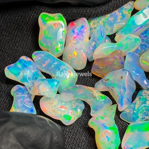 May include: A collection of iridescent opal gemstones in various shapes and sizes. The opals display a range of colors, including blue, green, and flashes of red and yellow. The text "Rajiyajewels" is visible on the surface.