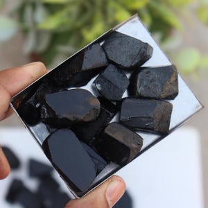May include: A collection of raw, black tourmaline stones displayed in a small, square metal tray. The stones have a rough, natural texture and varying shapes. The tray is held in a hand, with a blurred background.